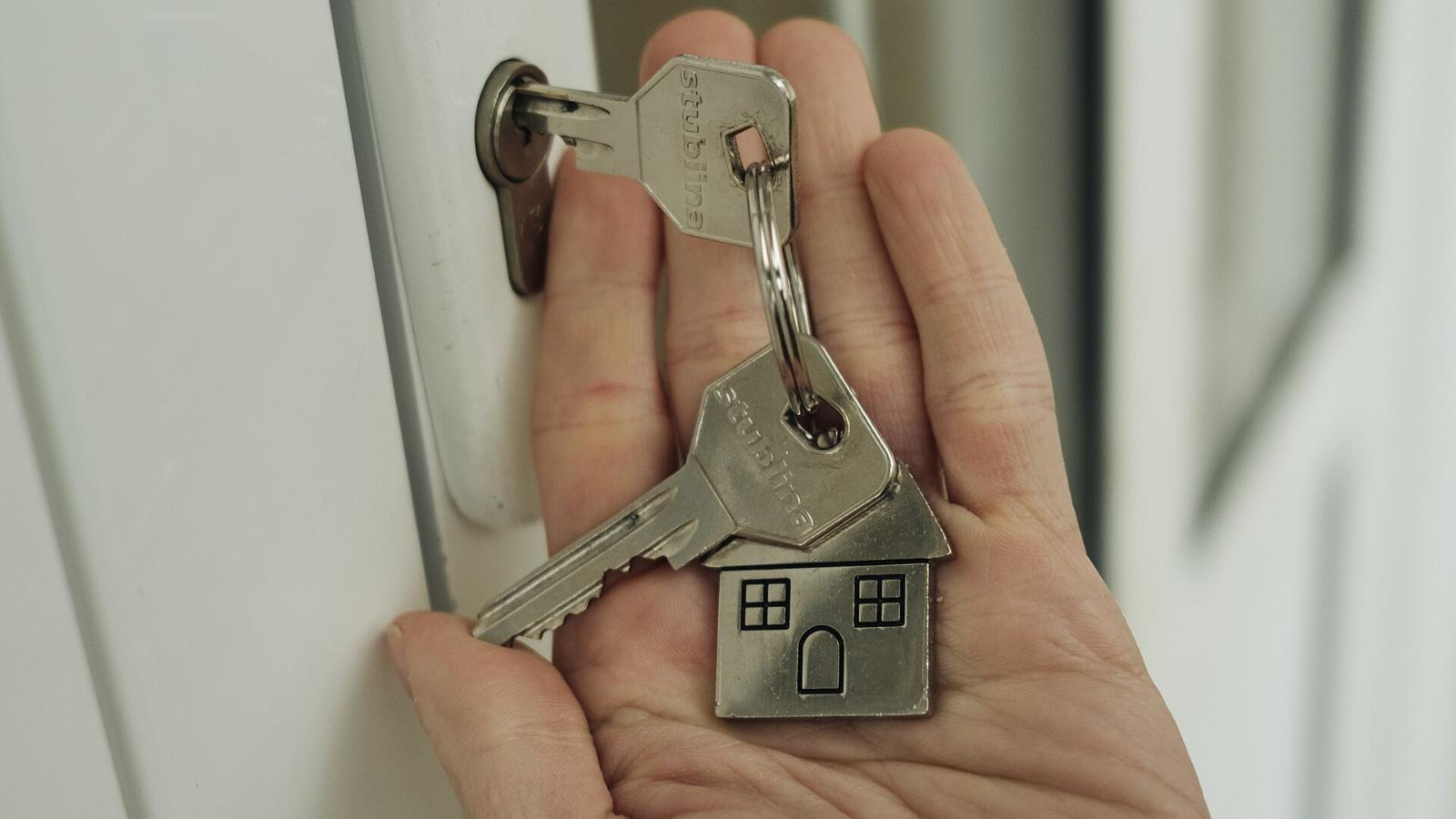 Una mano sostiene unas llaves con un llavero en forma de casa junto a una puerta